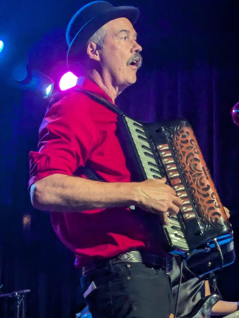 Krist Novoselic in a red shirt and black hat playing accordion on stage