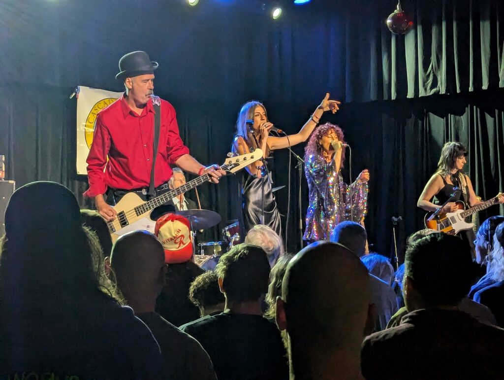 Krist Novoselic in a red shirt and black hat playing bass on stage with two singers and a guitarist, a Cascade Party banner hanging behind him
