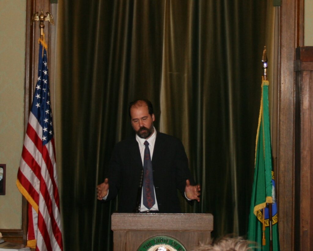 Krist Novoselic in a dark suit and tie speaking at a podium bearing the Washington state seal, between a U.S. flag and the Washington state flag
