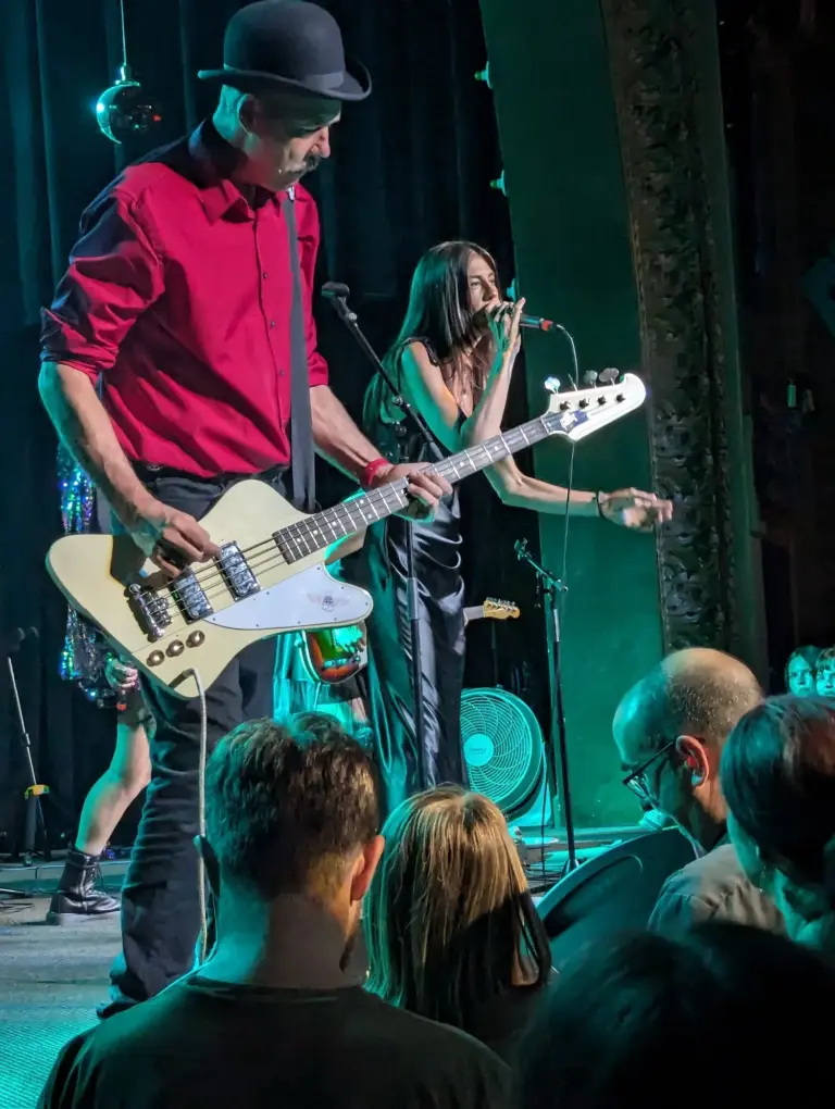 Krist Novoselic in a red shirt and bowler hat playing a cream Thunderbird-style bass on stage, with singer Jillian Raye behind him, at the Spanish Ballroom in Tacoma, Washington